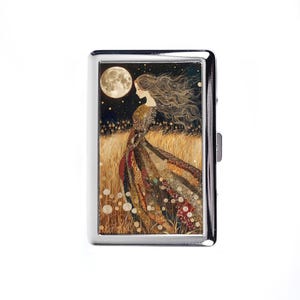 May include: A silver metal cigarette case featuring an illustration of a woman in a flowing dress standing in a field under a full moon. The artwork has a warm color palette with gold, brown, and red tones.