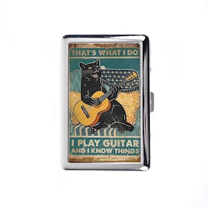 Op de afbeelding: Een zilveren metalen sigarettenetui met een zwarte kat die gitaar speelt. De etui heeft een vintage ontwerp met de tekst "THAT'S WHAT I DO I PLAY GUITAR AND I KNOW THINGS" in een verweerde lettertype. De kat staat op een patroonachtergrond.