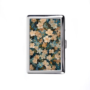 May include: A silver cigarette case with a floral design. The case features a pattern of cream, blue, and green flowers against a dark background. The case has a rectangular shape with a hinged opening and a metallic finish.