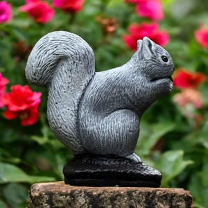 Puede incluir: Estatua de ardilla gris con cola tupida, posada sobre una base de piedra. La ardilla está sentada y sostiene algo en sus patas. El fondo presenta flores rojas y rosas.