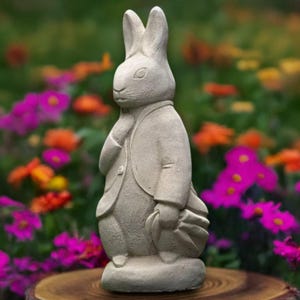 Puede incluir: Una estatua de conejo color piedra, vestida con una chaqueta y sosteniendo una bolsa, se encuentra sobre una superficie de madera. El conejo se encuentra sobre un fondo de flores coloridas, creando una encantadora escena de jard&iacute;n. La estatua es un objeto decorativo.