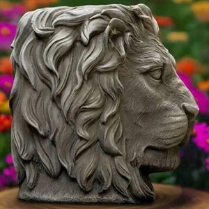 Puede incluir: Escultura de cabeza de león gris con detalles intrincados. La escultura muestra un perfil lateral de la cabeza del león, con una melena texturizada. El fondo está borroso, con flores coloridas.