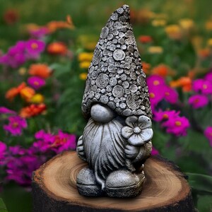 Puede incluir: Estatua de gnomo de jardín gris con sombrero y barba texturizados, sosteniendo una flor. El gnomo está sobre un tocón de madera, con un colorido jardín de flores al fondo.
