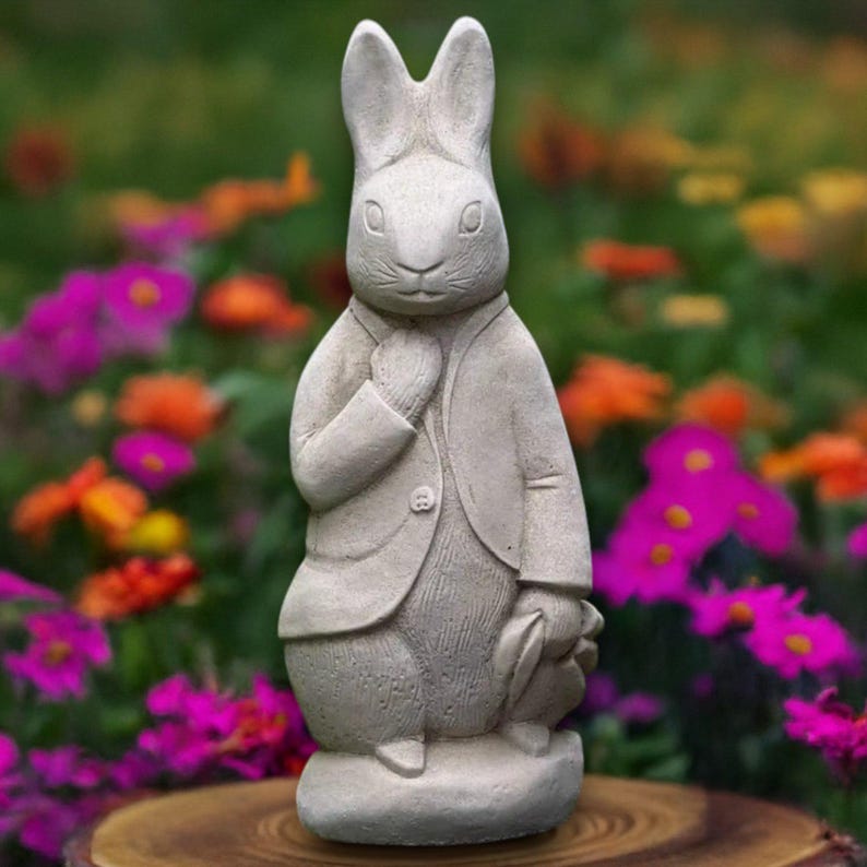 Puede incluir: Estatua de conejo de piedra gris claro, vestida con un traje y sosteniendo un sombrero. La estatua est&aacute; sobre una peque&ntilde;a base. El fondo presenta flores coloridas en tonos rosa, naranja y verde, creando una escena de jard&iacute;n.