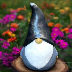 Puede incluir: Una figura de gnomo de jardín con un sombrero negro, barba blanca y cuerpo gris. El gnomo tiene una nariz amarilla y está sobre un fondo borroso de flores coloridas. El gnomo está sentado en un tocón de madera.