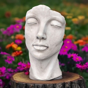 Könnte beinhalten: Ein hellgraues, skulptiertes Gesicht mit geschlossenen Augen, das auf einem Holzstumpf ruht. Die Skulptur hat einen ruhigen Ausdruck und einen Hintergrund aus bunten Blumen. Der Kopf ist eine Büste, wobei der obere Teil des Kopfes fehlt.