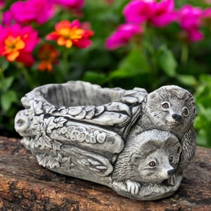 May include: A gray concrete planter shaped like a log, featuring two hedgehogs. The planter has a textured surface with leaf details. Bright pink and orange flowers are in the background.