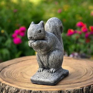 Puede incluir: Estatua de ardilla gris sosteniendo una nuez, sobre una superficie de madera. La ardilla tiene una textura de pelaje detallada y está sentada. El fondo presenta flores rosas borrosas y vegetación, creando un entorno natural.