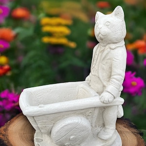 Puede incluir: Estatua de gato gris claro empujando una pequeña jardinera de carro. El gato lleva una chaqueta y pantalones. La jardinera tiene una abertura rectangular. El fondo es una vista borrosa de flores coloridas.