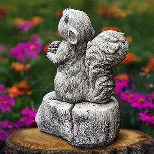 Puede incluir: Una estatua de ardilla gris sosteniendo una piña, posada sobre una base de piedra. La ardilla tiene detalles de pelaje y mira hacia arriba. La estatua está situada sobre un fondo borroso de flores coloridas, creando un encantador acento de jardín.