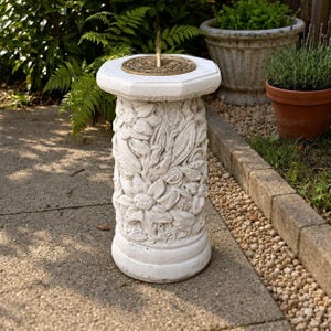 May include: A white concrete sundial featuring a cylindrical pedestal with relief carvings of flowers and leaves. The top surface is octagonal with a circular bronze-colored dial plate and a central vertical gnomon casting a shadow.