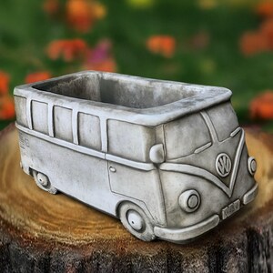 May include: A gray concrete planter shaped like a vintage Volkswagen bus. The planter has a rectangular opening for plants and features detailed molding of the bus's front, sides, and wheels. The VW logo is visible on the front.