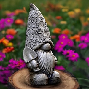 Puede incluir: Estatua de gnomo de jardín gris con una larga barba texturizada y un sombrero puntiagudo decorado con patrones circulares. El gnomo sostiene una pequeña pala y está de pie sobre un tocón de madera, con un colorido jardín de flores al fondo.