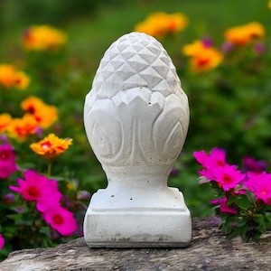 Op de afbeelding: Lichtgrijs betonnen tuinornament in de vorm van een dennenappel. Het ornament heeft een textuuroppervlak en staat op een vierkante basis. De achtergrond toont kleurrijke bloemen in geel, oranje en roze.