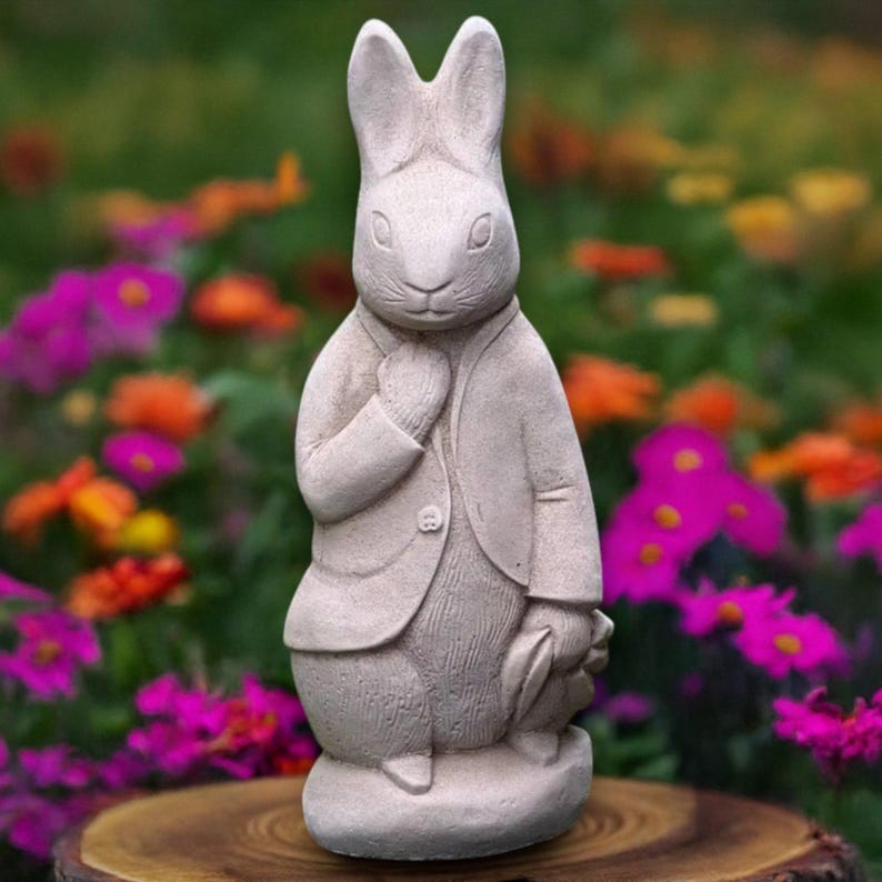 Puede incluir: Una estatua de conejo color piedra, vestida con una chaqueta, se encuentra sobre una superficie de madera. El conejo se lleva la mano al pecho, con una peque&ntilde;a hoja en la otra mano. El fondo presenta un jard&iacute;n colorido con varias flores.