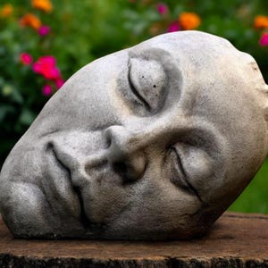 Könnte beinhalten: Eine verwitterte, graue Steinskulptur eines menschlichen Gesichts mit geschlossenen Augen. Die Skulptur ruht auf einer Holzoberfläche, mit einem verschwommenen Hintergrund aus grünem Laub und bunten Blumen. Das Gesicht hat detaillierte Merkmale und einen ruhigen Ausdruck.