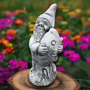 Puede incluir: Estatua de gnomo de jardín gris sosteniendo un pez. El gnomo tiene una larga barba blanca y un gorro puntiagudo. El pez tiene un ojo grande y escamas. La estatua está sobre una base de madera, con un jardín de flores coloridas al fondo.
