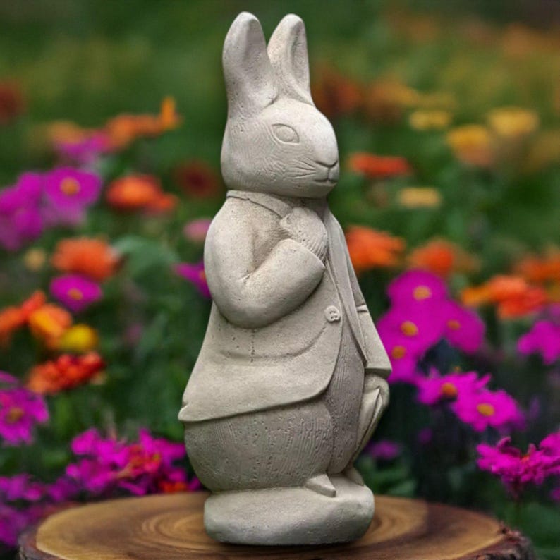 Puede incluir: Estatua de conejo color piedra, vestida con traje y corbata, de pie sobre una superficie de madera. El conejo tiene orejas largas y est&aacute; posando como si mirara hacia un lado. El fondo es un jard&iacute;n colorido con flores.