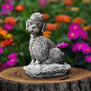 Puede incluir: Una estatua detallada de un caniche gris sentada sobre un cojín decorativo con un diseño de rosas. El perro tiene un pelaje texturizado y lleva un collar. La estatua está colocada sobre un tocón de árbol de madera, con un jardín de flores coloridas en el fondo.