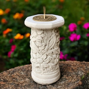 May include: A white concrete sundial with a floral design on the column. The sundial has a bronze-colored face and gnomon. The sundial is sitting on a rock with a blurred background of colorful flowers.