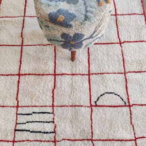 Puede incluir: Una alfombra color crema con un patrón de cuadrícula roja y formas negras abstractas. Un otomán con estampado floral con patas de madera descansa sobre la alfombra. La alfombra tiene una apariencia texturizada y afelpada.