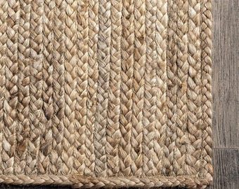 May include: Close-up of a natural jute rug with a braided texture. The rug's neutral beige color and thick weave create a rustic, textured appearance. The rug is placed on a wooden floor.