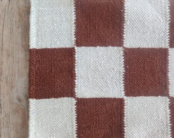 May include: A close-up of a woven rug with a checkerboard pattern. The rug features alternating squares of brown and off-white. The texture of the rug appears to be slightly rough, with visible fibres. The rug is placed on a wooden surface.
