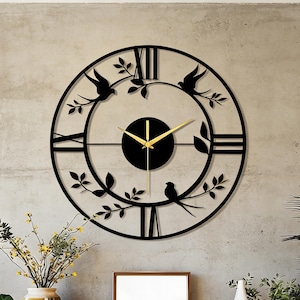 May include: A black metal wall clock with a circular design. The clock features Roman numerals, gold hands, and decorative bird and leaf accents. The clock is mounted on a gray concrete wall above a wooden table with decorative items.