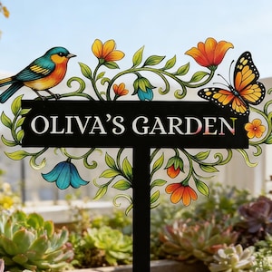 May include: A black metal garden sign with the text "OLIVA'S GARDEN". The sign is decorated with a colorful bird, butterfly, flowers, and foliage. The sign is placed in a garden with various plants and succulents.