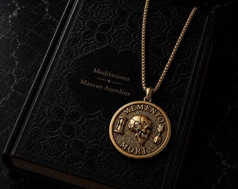 Könnte beinhalten: Eine goldfarbene, kreisförmige Anhängerkette mit einem Relief aus Totenkopf, Sanduhr und Pfeil sowie dem Text MEMENTO MORI, die auf einem schwarzen Hardcover-Buch mit dem Titel Meditations von Marcus Aurelius und geprägtem Blumenmuster liegt.