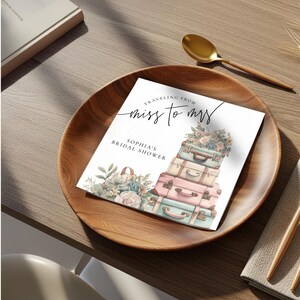 May include: A square invitation card with the text "Traveling from miss to mrs" and "Sophia's Bridal Shower". The card features a stack of pastel-colored suitcases and floral arrangements, set on a wooden plate.