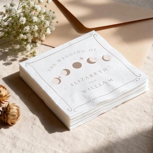 May include: A stack of white paper napkins with a decorative border and text that reads "THE WEDDING OF ELIZABETH and WILLIAM". The design includes moon phases and stars in a neutral color palette, suitable for a wedding.