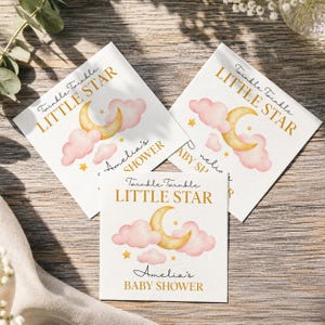 May include: Three square white paper napkins featuring a watercolor illustration of a yellow crescent moon, pink clouds, and small gold stars. The text reads Twinkle Twinkle LITTLE STAR and Amelia's BABY SHOWER in gold and black script and block fonts.