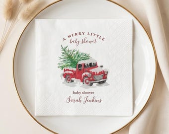 May include: A white paper napkin featuring a watercolour illustration of a red truck carrying a green Christmas tree. The text reads A MERRY LITTLE baby shower and Sarah Jenkins in dark red script and serif fonts, centred on the square napkin.