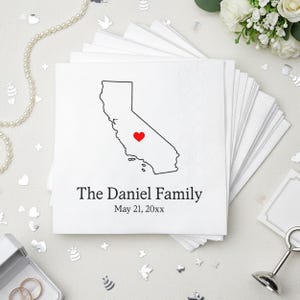 May include: White paper napkins with a black outline of California and a red heart. The text "The Daniel Family" and the date "May 21, 20xx" are printed below the map. The napkins are stacked in a neat pile.