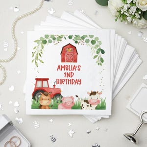 May include: White square napkins with a farm-themed design. The design includes a red barn, tractor, and cartoon animals. The text "AMELIA'S 2ND BIRTHDAY" is printed in red. Green foliage frames the top of the design.