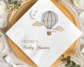 May include: White square paper napkin featuring a watercolour illustration of a hot air balloon, clouds, moon, and stars. The text "HENRY'S Baby Shower" is printed in brown script. The napkin is on a gold-toned plate, surrounded by decorations.