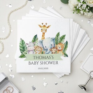 May include: White square napkins with a watercolor illustration of jungle animals, including a giraffe, elephant, lion, and monkey. The napkins have the text "THOMAS'S BABY SHOWER" and the date 05.02.20XX printed on them. The napkins are stacked.