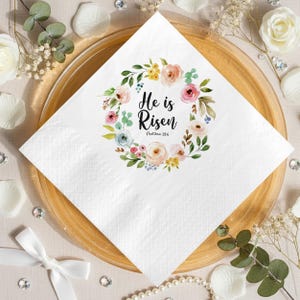 May include: White square paper napkin with a floral wreath design. The wreath features pink, peach, yellow, and blue flowers with green leaves. The text "He is Risen" is in the center, with "Matthew 28:6" below.