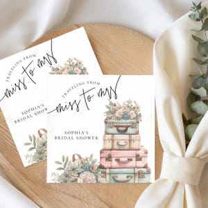 May include: Bridal shower invitation featuring a stack of vintage suitcases in pastel colors, topped with flowers. The text reads "Traveling from Miss to Mrs" and "Sophia's Bridal Shower". The design includes floral accents.