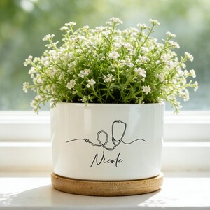 May include: A white ceramic planter with a stethoscope design and the name "Nicole" in black script. The planter holds a small green plant with white flowers and sits on a wooden coaster. The background is a blurred view of green foliage.