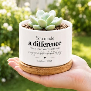 May include: A white ceramic cylindrical planter containing a light green succulent. The planter features black text reading: You made a difference more than words can say may your future be full of joy, with a heart icon, Sophia, and 2026. It sits on a wooden base.