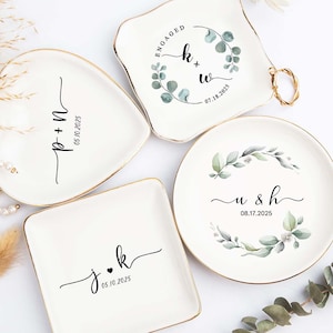 Puede incluir: Conjunto de platos de cerámica blanca para anillos con borde dorado. Cada plato presenta una forma y un diseño únicos, incluyendo un corazón, un cuadrado y un círculo. Los platos están decorados con iniciales, fechas e ilustraciones botánicas.