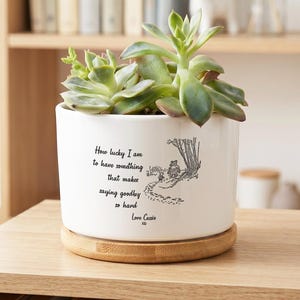May include: A white ceramic cylindrical planter with a bamboo saucer base containing a green succulent plant. The planter features a black line drawing of two characters walking on a path with the text: How lucky I am to have something that makes saying goodbye so hard. Love Cassie xx.