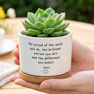 May include: A white ceramic cylindrical planter containing a green succulent plant. The planter features black printed text that reads: Be proud of the work you do, the brilliant person you are and the difference you make! Emma! with a puzzle piece icon.