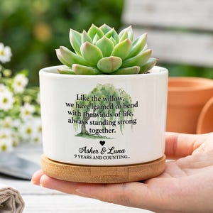 Könnte beinhalten: Ein weißer Keramikübertopf mit Bambusuntersetzer und einer grünen Sukkulente. Der Übertopf zeigt eine Aquarell-Illustration eines Weidenbaums und den Text: Like the willow, we have learned to bend with the winds of life, always standing strong together. Asher & Luna 9 YEARS AND COUNTING.