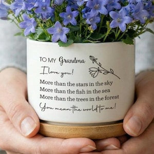 May include: A white ceramic flower pot filled with vibrant blue flowers, held in someone's hands. The pot has the text "TO MY Grandma I love you!" and other loving phrases. The pot sits on a wooden base, creating a thoughtful gift.