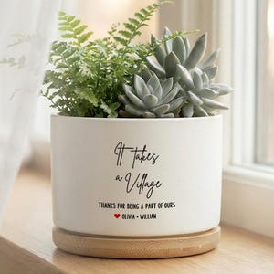 May include: A white ceramic plant pot with the text "It takes a Village" and a bamboo base. The pot contains green plants, including ferns and succulents. The text also includes "Thanks for being a part of ours" and a heart symbol.