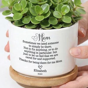 May include: A white ceramic planter with a green succulent plant, on a round wooden coaster. The planter has the word "Mom" and a sentimental message printed on it. The text includes a name and the word "xoxo".
