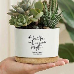 Op de afbeelding: Een witte keramische plantenbak met een bamboe basis, gevuld met vetplanten en cactussen. De plantenbak heeft de tekst "A beautiful soul is never forgotten" en de naam "EMMA SWAN 1954-2025".